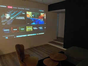 A room with a large screen displaying a Canal+ interface, a wooden floor, a brown armchair, and a small round table.