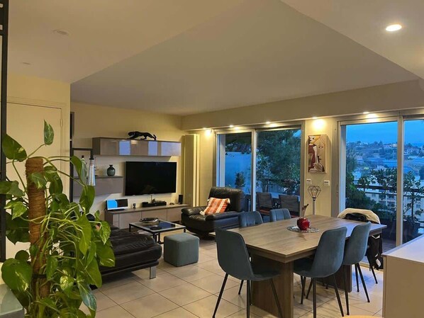 A modern living room with a flat-screen TV, a sofa, a dining table with chairs, and a large window offering a city view.