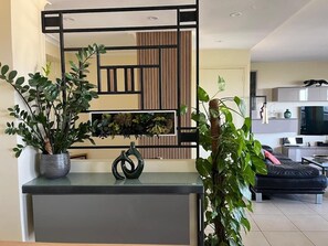 Un salon moderne avec un canapé, une table basse et un téléviseur. Un panneau décoratif orne le mur, et une plante en pot se trouve sur la gauche.