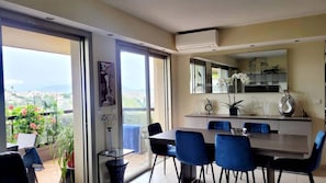 Un comedor moderno con una mesa grande y sillas azules, una cocina con mostrador y armarios, y un balcón con vistas a la ciudad.