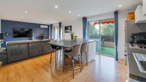 A modern living room with a sofa, a dining table with chairs, a flat-screen TV mounted on the wall, and a kitchen area with a stove. - Comfortable Apartment with large sunny terrace (Niza)