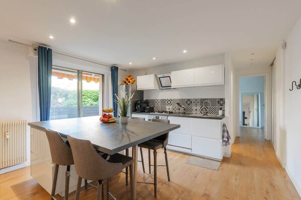 A modern kitchen with a dining area, white cabinets, a tiled backsplash, and a view through the window.