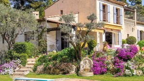 Una casa de dos pisos con piscina, rodeada de abundante vegetación y plantas florecientes.