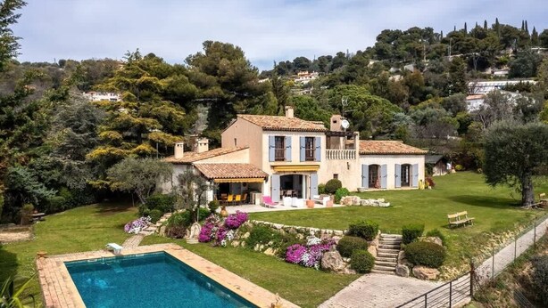 Una casa grande con piscina, rodeada por un jardín cuidado y un paisaje montañoso.