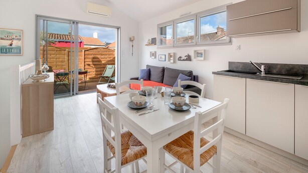 Una cocina moderna con armarios blancos, una mesa de comedor dispuesta con platos y cubiertos, y vista a un edificio de tejados rojos a través de una puerta corrediza de vidrio.