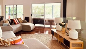 Un salon moderne avec un canapé, une table basse et un meuble pour la télévision. Une grande fenêtre offre une vue sur la verdure à l’extérieur.