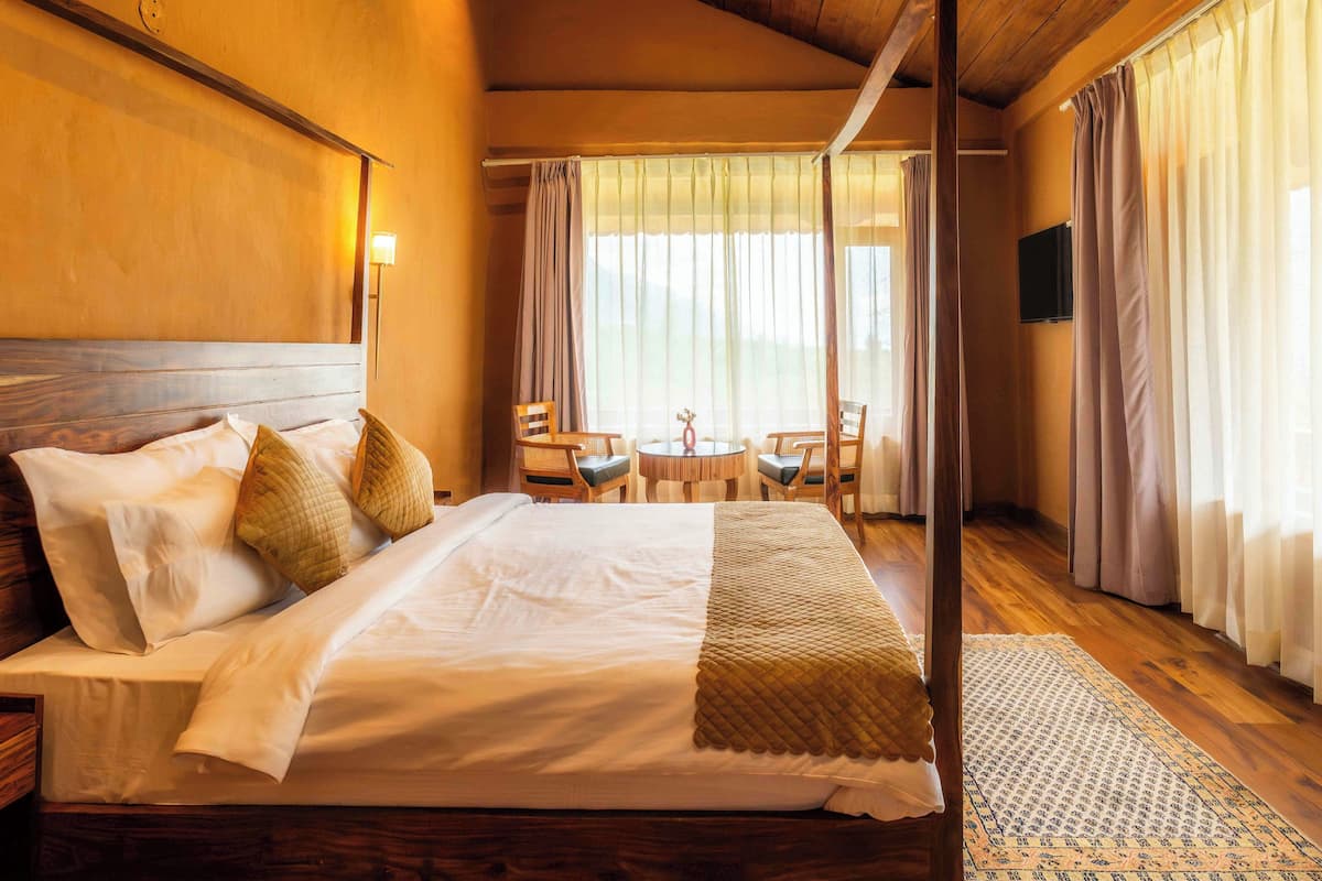 Habitación de hotel con una cama grande, cabecera de madera, mesita de noche, una silla, una pequeña mesa con lámpara y un televisor en la pared.