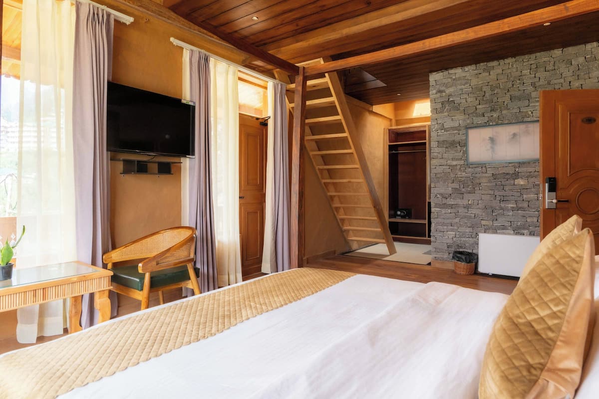 Un dormitorio con cama, escritorio, silla, televisor, escalera y pared de piedra.