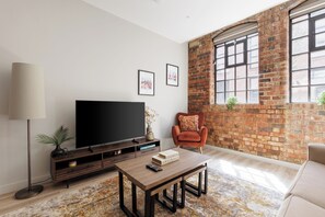 A living room with a flat-screen TV, a coffee table, a sofa, a brick wall, and large windows.