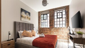 A modern bedroom with a brick wall, a large bed with a red blanket, a nightstand, a desk with a plant, and a TV. - The Brickworks - Grade II Boutique Stay, 2 Bed. (Birmingham)