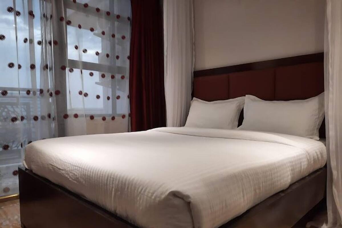 A neatly made bed with a dark headboard, white bedding, and sheer curtains with a floral pattern.