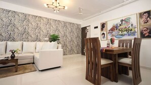 A living room with a white sofa, a dining table with wooden chairs, a ceiling fan, and a wall with Bollywood-themed artwork. - 2 BHK HomeSuite (Gagal Home) (Mumbai)