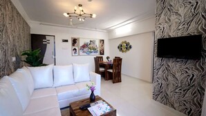 A modern living room with a white sofa, a wooden coffee table, and a dining area with a wooden table and chairs. There is a wall-mounted TV and a decorative wall hanging. - 2 BHK HomeSuite (Gagal Home) (Mumbai)
