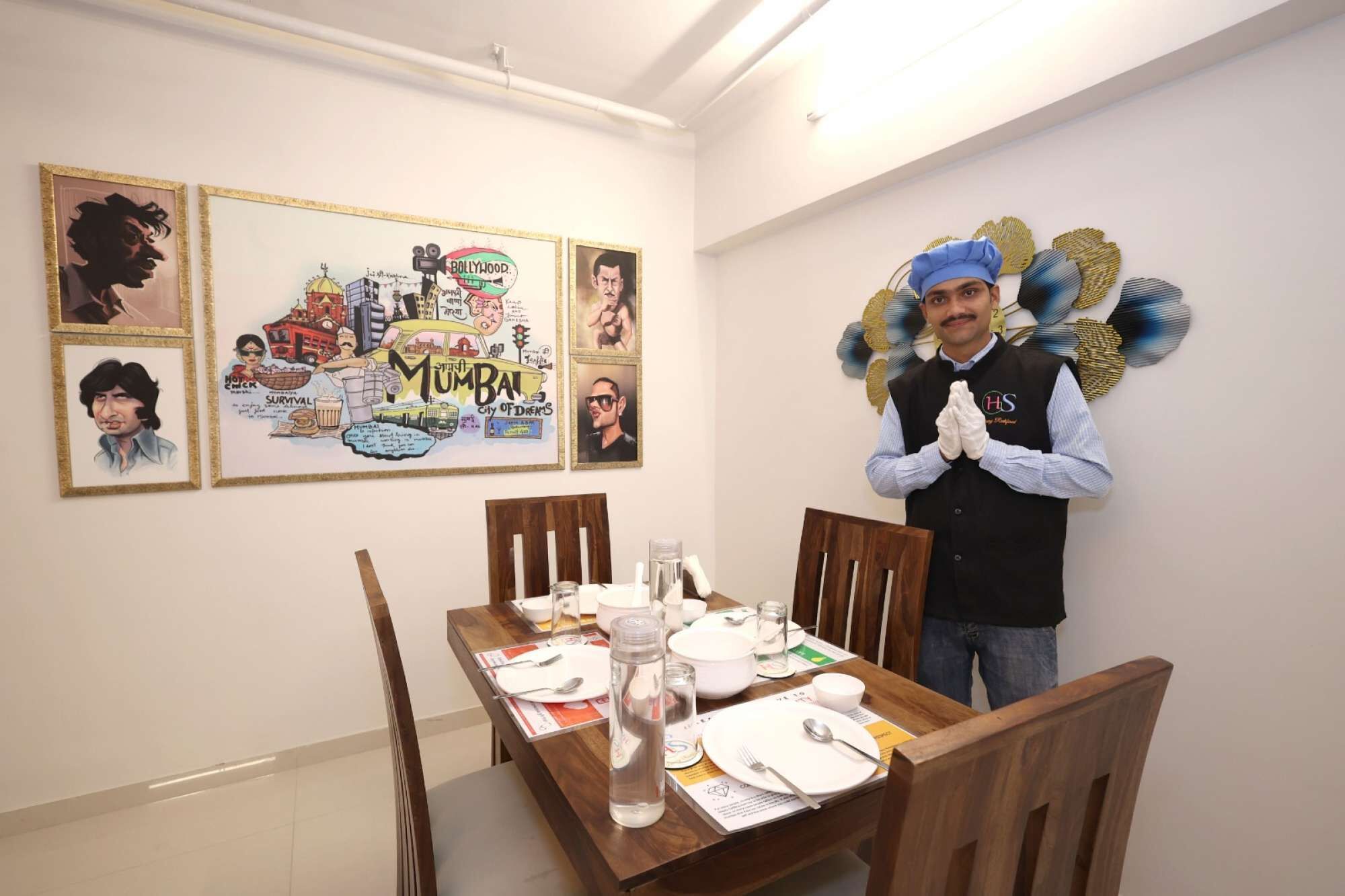 A dining area with a wooden table set for four, a decorative wall with framed pictures, and a person standing in the background.