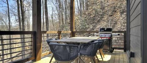 An outdoor deck with a table and chairs, a grill, and a view of a forested area.