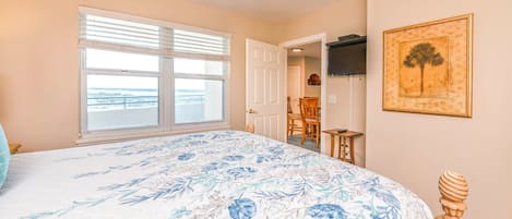Un dormitorio con una cama grande, un ventilador de techo, una ventana con persianas, una puerta que da a un comedor y un cuadro en la pared.