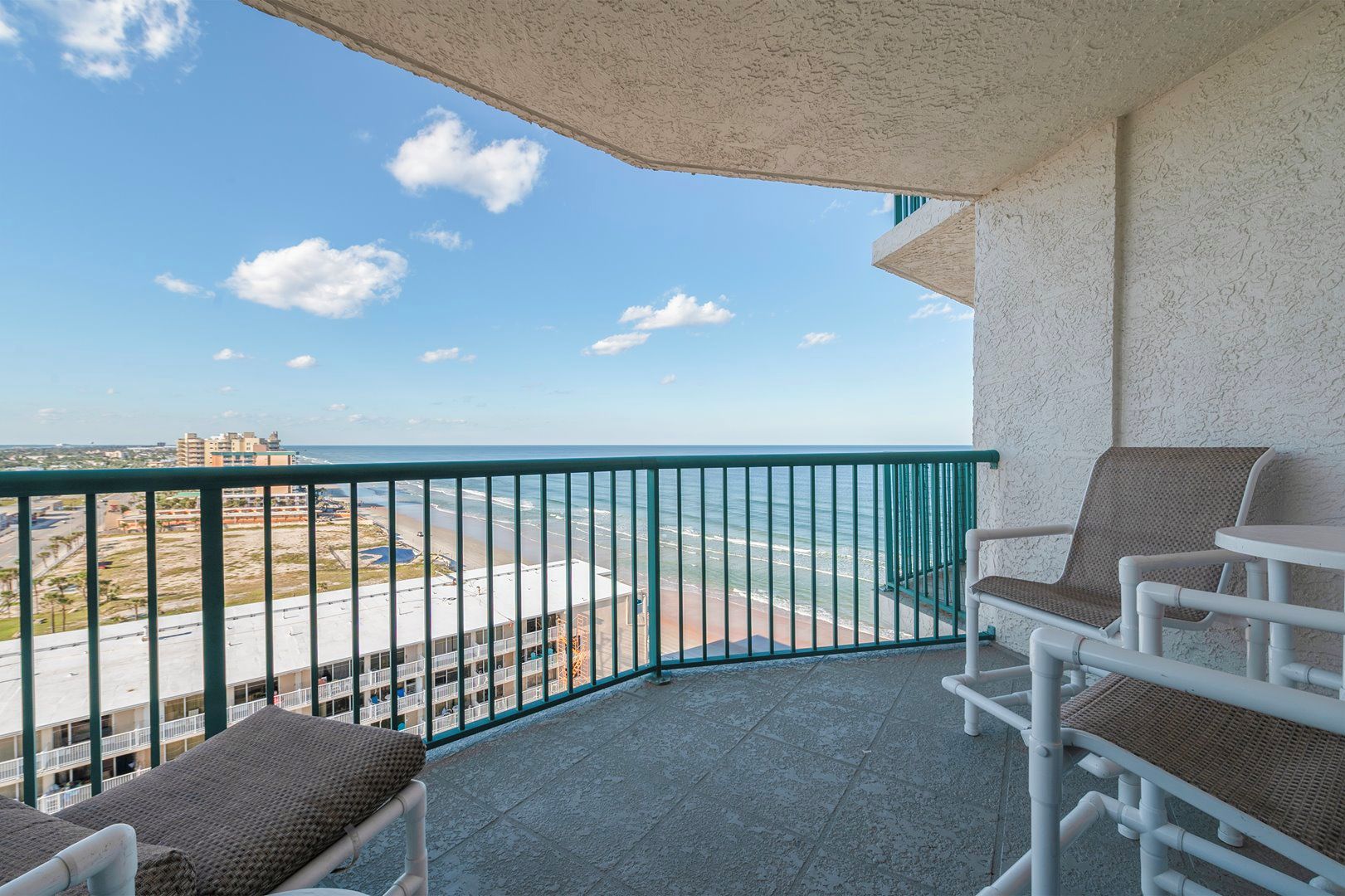 A balcony with a view of the beach and ocean, featuring two wicker chairs and a small table.