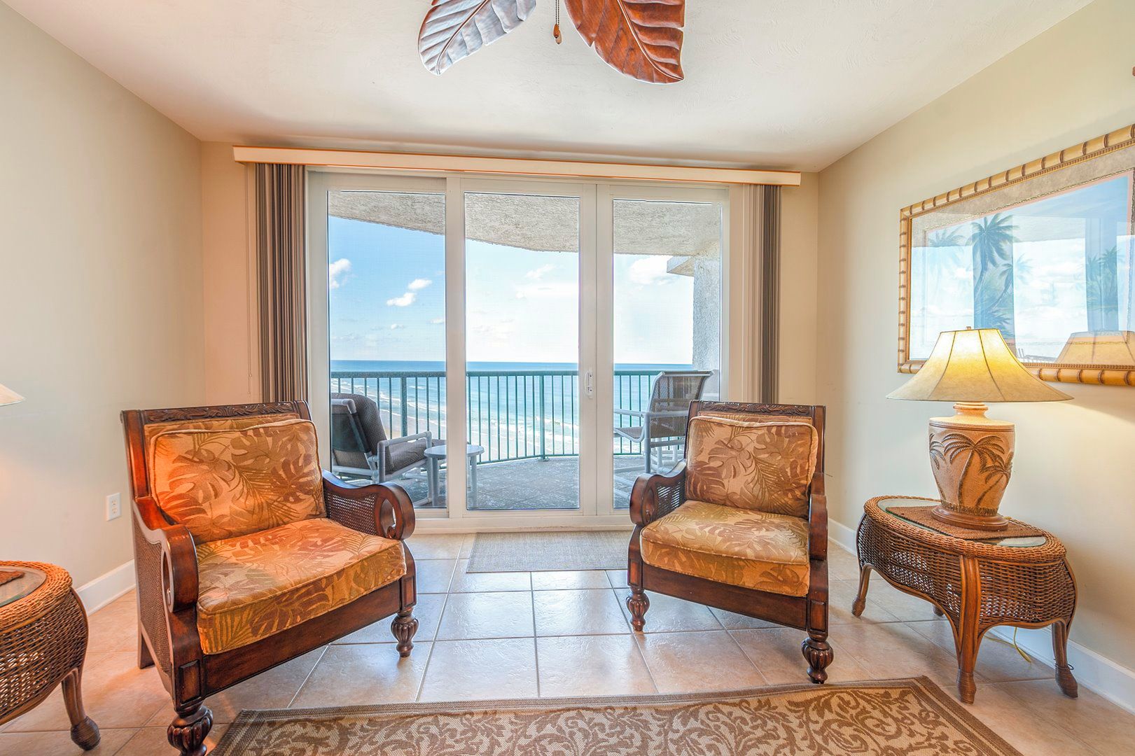A room with two upholstered chairs, a small table with a lamp, and a view of the ocean through a sliding glass door.