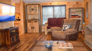 A cozy living room with a wooden floor, a flat-screen TV, a leather armchair, a red sofa, and a wooden coffee table with a board game. - Sleeps 6・Cozy Cabin・Spacious Outdoor・View・Firepit (Sevierville)
