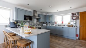 A modern kitchen with a large island, bar stools, and a view of the living area. - Beach Front 5 Star Dog Friendly Home with Amazing Sea Views & Outdoor Kitchen (Portreath)