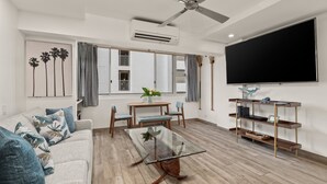 Een moderne woonkamer met een glazen salontafel, een bank met gekleurde kussens, een eettafel met stoelen, een grote flatscreen televisie aan de muur en een plafondventilator.