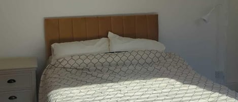 A bed with a patterned bedspread and two pillows, a wooden headboard, and a bedside table with drawers.