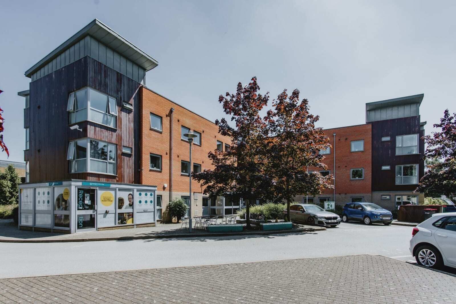 Un edificio moderno con recepción, una pequeña zona de estar al aire libre y un estacionamiento con coches.