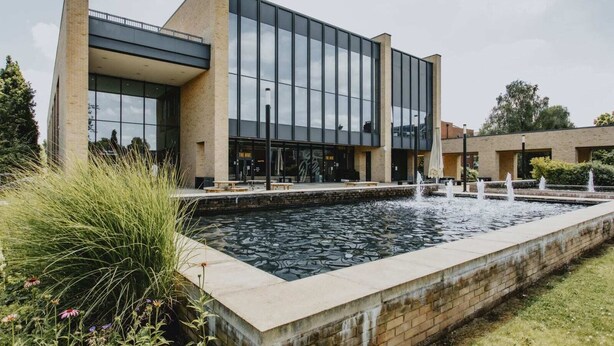 A modern building with a large glass facade, a brick base, and a rectangular water feature in front. - Bonington Student Village - Campus Accommodation (Sutton Bonington)