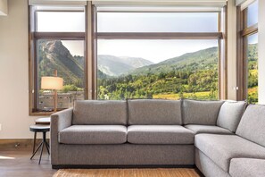 A modern living room with a large sectional sofa, a small table, and a lamp, with a scenic mountain view through large windows. - New Listing! Ski In/Ski Out | Riverfront Views | Spa (Avon)