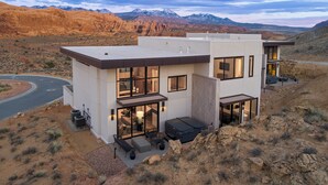 A modern two-story house with large windows, a patio with outdoor furniture, and a view of mountains in the background. - Slickrock Luxury Retreat | Panoramic Views | Min to Trails and Ntl Parks (Moab)
