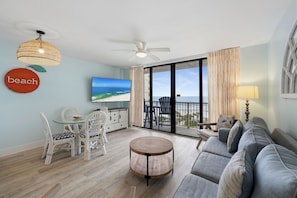 Una sala de estar moderna con un sofá, una mesa redonda de centro, una mesa de comedor con sillas, un televisor y un balcón con vista a la playa.