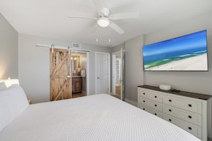 Une chambre à coucher avec un lit, un ventilateur de plafond, un téléviseur fixé au mur et une porte en bois donnant accès à une salle de bain.