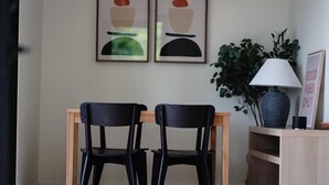 Une pièce avec une table en bois et deux chaises noires, une plante en pot, une lampe et deux œuvres d’art abstraites encadrées accrochées au mur.