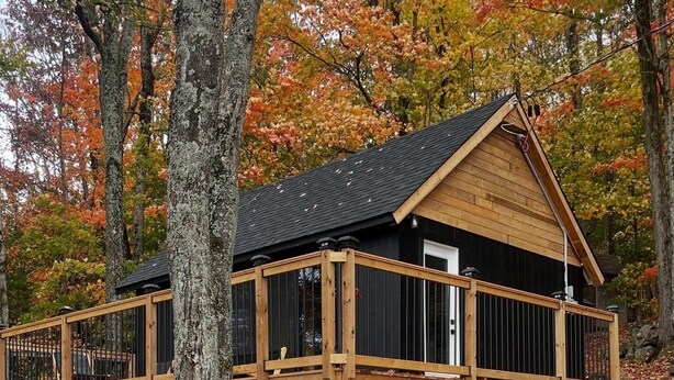 Une cabane dotée d’une terrasse en bois et d’un toit noir, entourée du feuillage automnal.