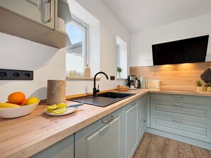A modern kitchen with a wooden countertop, a black sink, and light blue cabinets. There is a window with blinds, a wall-mounted TV, and a bowl of fruit on the counter. - Haus Hygge - with sauna and fireplace 1700 - Haus Hygge Am Kurpark with sauna and fireplace 1700 (Dahme)