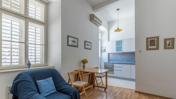 A living room with a blue sofa, a wooden dining table with chairs, a vase with flowers, and a kitchen in the background. - Central Cathedral Apartment (Kotor)