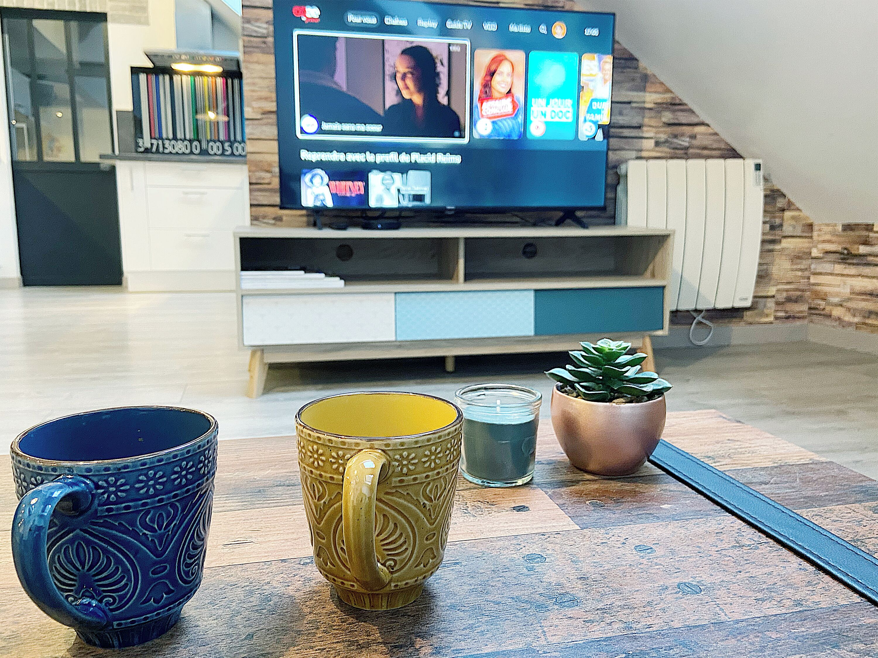 A living room with a flat-screen TV, two decorative mugs, a candle, and a potted plant.