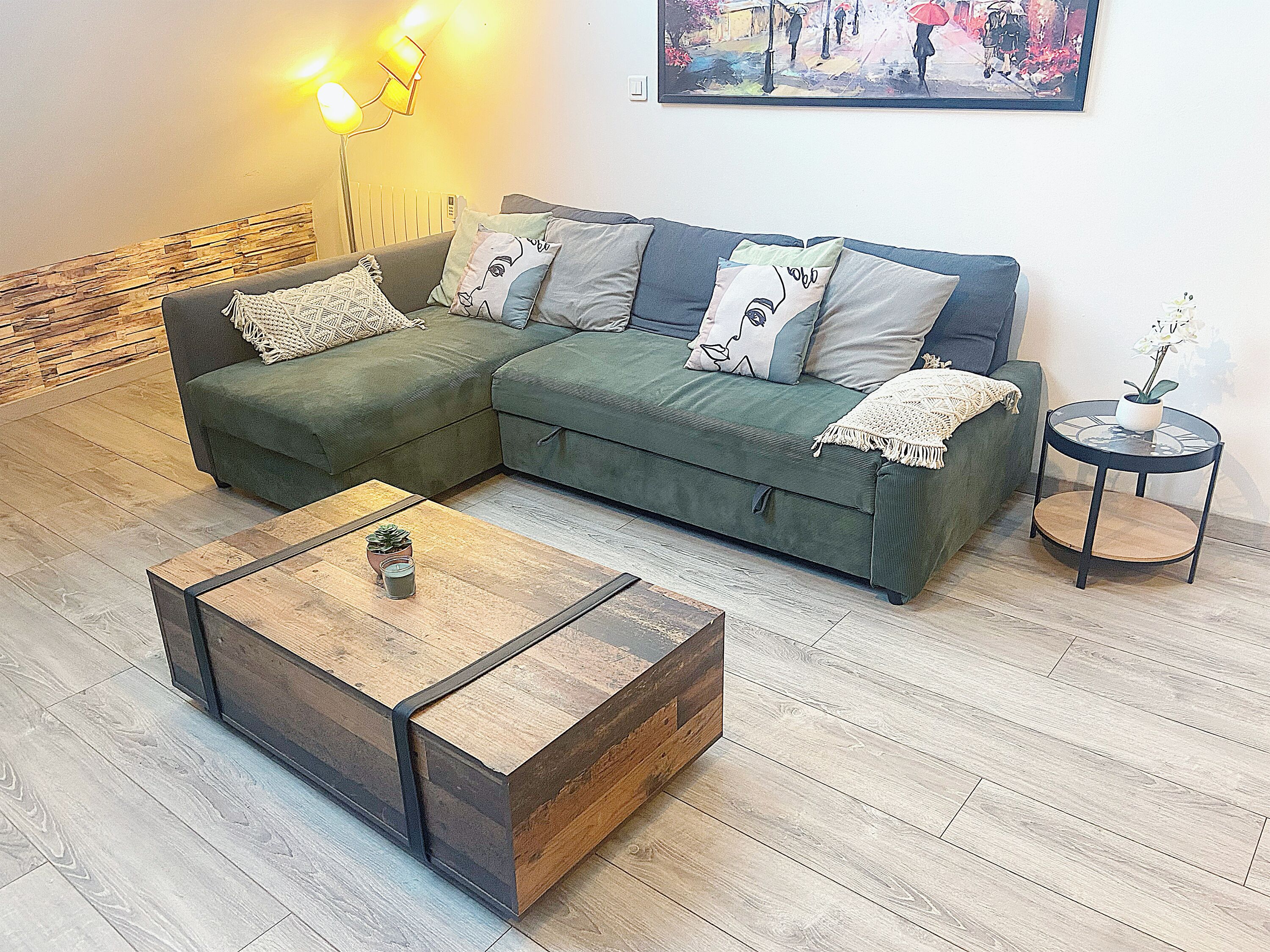 Una sala de estar con un sofá modular, una mesa de centro, una mesita de noche con una planta y un cuadro en la pared.
