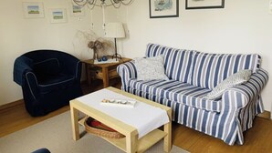 Een woonkamer met een gestreepte bank, een donkere fauteuil, een houten salontafel, een kroonluchter en lijstwerken aan de muur.