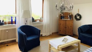 Een gezellige woonkamer met een blauwe fauteuil, een houten kast en een tafel met een witte tafelkleed.