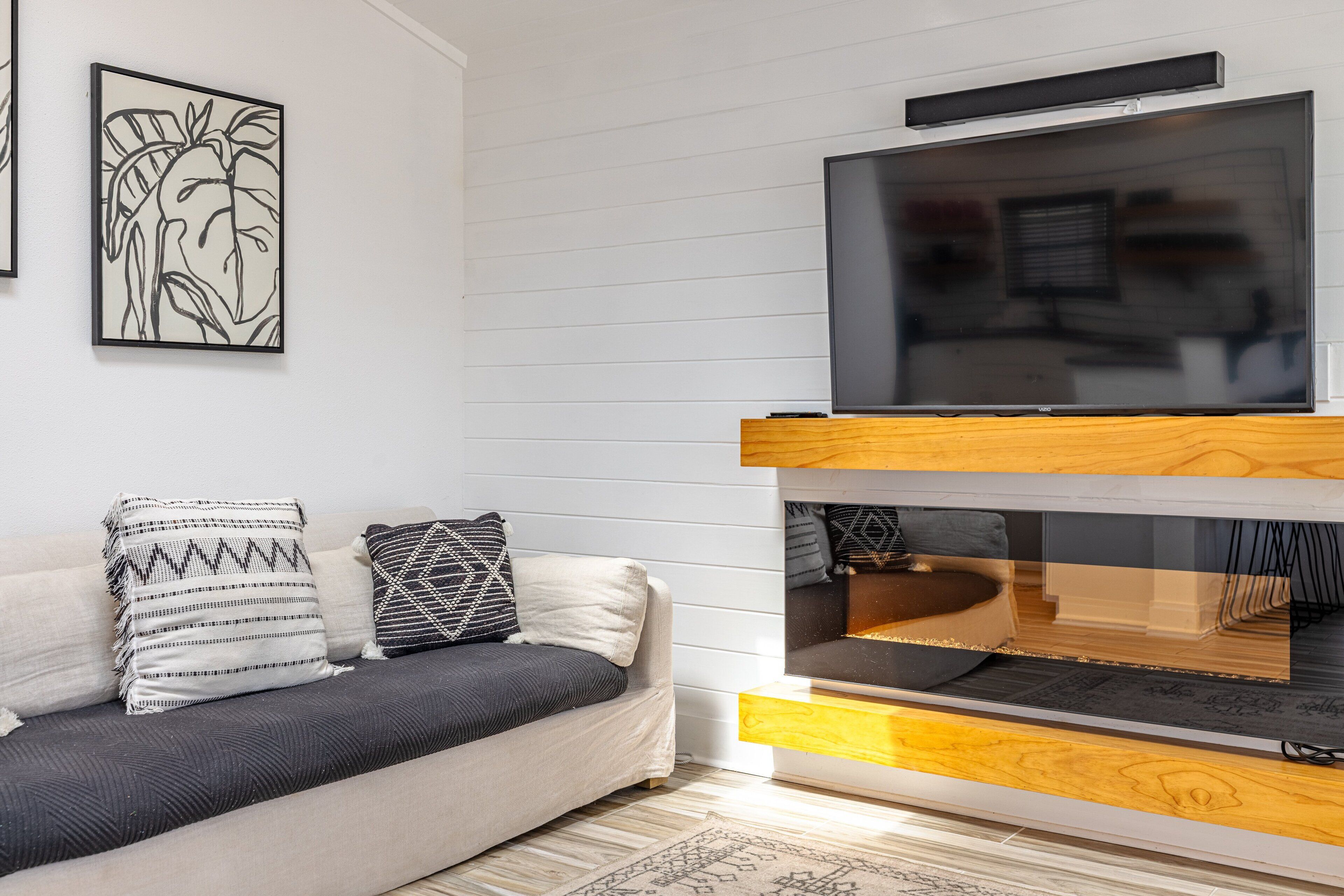 Ein modernes Wohnzimmer mit grauem Sofa, gemusterten Kissen, einem Flachbildfernseher auf einem Holzboden und einem gerahmten Kunstwerk an der Wand.