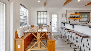 Une cuisine moderne avec une table en bois et des tabourets de bar, un coin salon avec un banc, et une vue sur l’extérieur grâce à de grandes fenêtres.