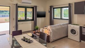 A compact living space with a sofa, a washing machine, a dining table with chairs, a TV mounted on the wall, and large windows with curtains. - Casa Barbara (Chania)
