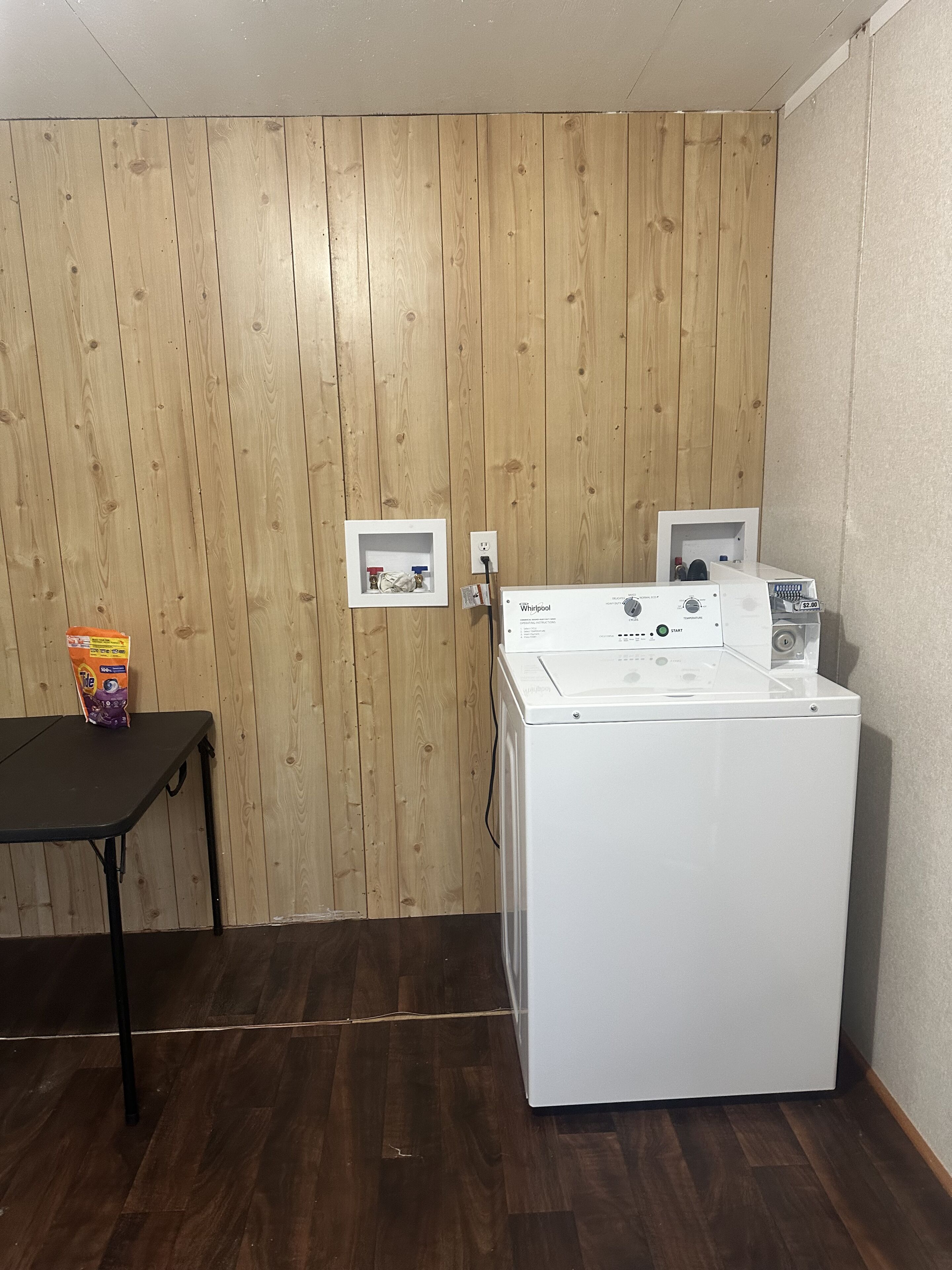 Una sala de bugaderia amb una rentadora i assecadora Whirlpool blanques, parets de fusta, una taula petita amb una bossa de detergent i una presa elèctrica.