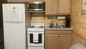 Cocina compacta con gabinetes de madera, refrigerador blanco, microondas, estufa y un cubo de basura.