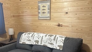 Un sofá gris con una manta estampada, una ilustración enmarcada de un pez y un reloj en una pared de madera.