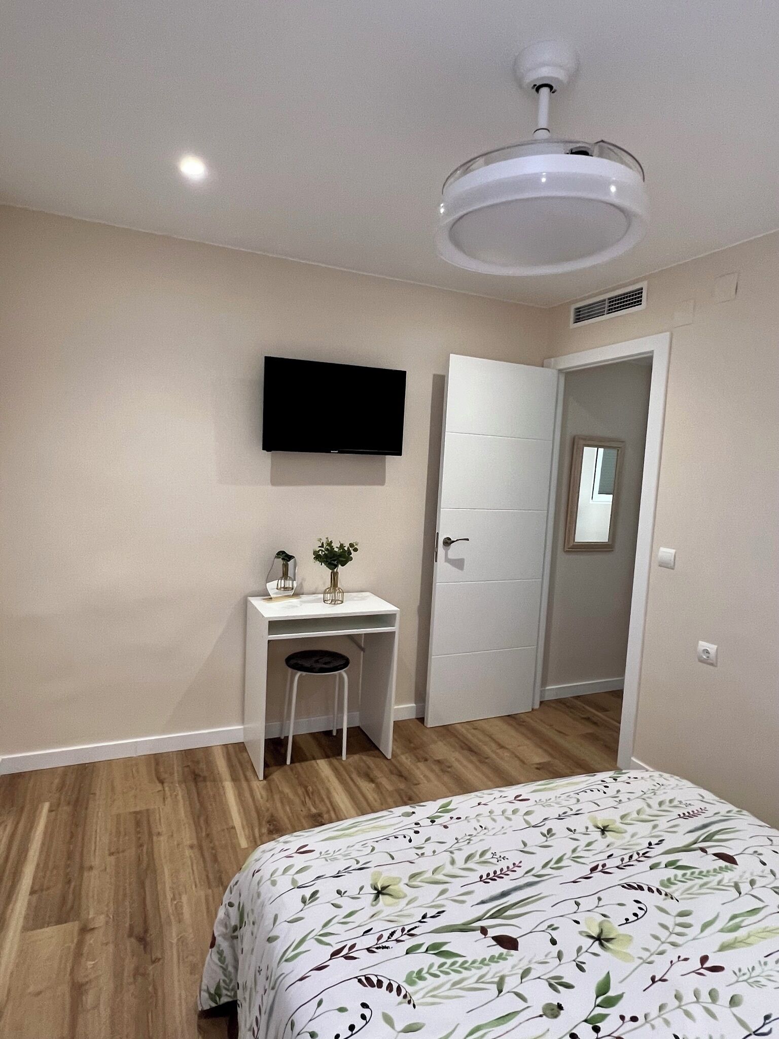 Une chambre à coucher avec un lit, un bureau et une chaise, un téléviseur fixé au mur, et une porte donnant sur une autre pièce.