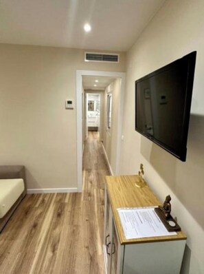 Un pasillo con suelo de madera, una cama, un escritorio con televisor y una rejilla en la pared.