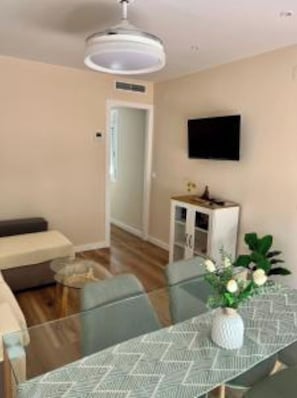 Una sala de estar moderna con una mesa de comedor de vidrio, un sofá, un armario blanco y un televisor de pantalla plana montado en la pared.
