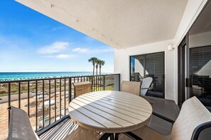 A balcony with a round table and chairs, overlooking a beach with palm trees and umbrellas. - Emerald Towers #205 OnTheGulf*LotsOfResortAmenites (DESTIN)
